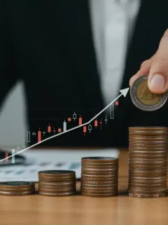 Hand placing coin on stacked coins with rising graph and house, symbolizing interest rate impact.